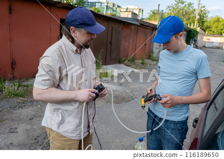 Dad and teenage son work together on the street near the garage. Outdoor activity. Family spend time. Helping and teach. Repairing a hose and pump for hand washing a car from a plastic bottle 116189650