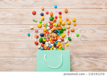 shopping paper gift bag in corner full of assorted traditional candies falling out on colored background with copy space. Happy Holidays sale concept shopping paper gift bag in corner full of assorted traditional candies falling out on colored background with copy space. Happy Holidays sale concept 116191259