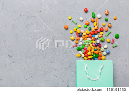 shopping paper gift bag in corner full of assorted traditional candies falling out on colored background with copy space. Happy Holidays sale concept shopping paper gift bag in corner full of assorted traditional candies falling out on colored background with copy space. Happy Holidays sale concept 116191260