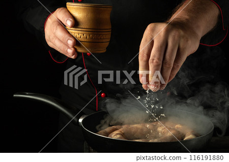 Chef salting meat sausage while frying it in a pan. Low key concept of cooking sausage in a tavern kitchen Chef salting meat sausage while frying it in a pan. Low key concept of cooking sausage in a tavern kitchen 116191880