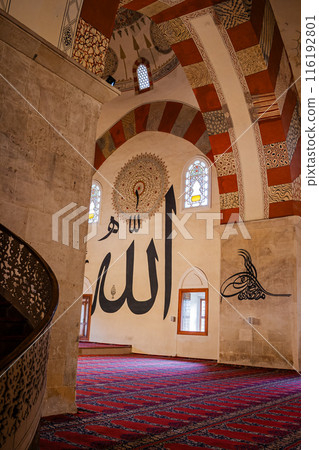 Interior of a mosque with large Arabic calligraphy on the walls and pillars with people are seen walking and praying on a red patterned carpet, in Edirne, Turkey. Interior of a mosque with large Arabic calligraphy on the walls and pillars with people are seen walking and praying on a red patterned carpet, in Edirne, Turkey. 116192801