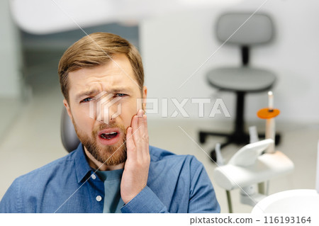 Sad man sitting in dental chair suffering from acute tooth pain, rubbing his cheek, close up Sad man sitting in dental chair suffering from acute tooth pain, rubbing his cheek, close up 116193164