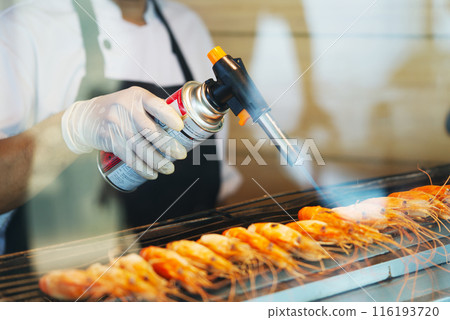 Close up hand with rubber glove holding hand held food burner burning prawns on charcoal stove, space for copy and design, landscape image. 116193720