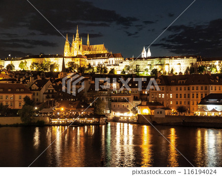Prague Castle illuminated at night, with the Vltava River reflecting the city lights. Buildings and spires are visible against the dark sky. 116194024