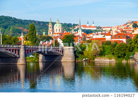 A stone bridge spans the Vltava River in Prague, Czechia. Red-roofed buildings line the hills in the background, with a church spire visible. 116194027