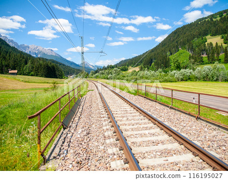 In the Dolomites, Italy, a set of train tracks runs through a scenic valley surrounded by green fields, forests, and snow-capped mountains under a bright blue sky. In the Dolomites, Italy, a set of train tracks runs through a scenic valley surrounded by green fields, forests, and snow-capped mountains under a bright blue sky. 116195027