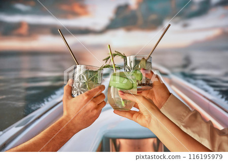 Group of people, friends relaxing on yacht in the evening, hands clinking glasses with cocktails, enjoying tranquil nature and sunset. Group of people, friends relaxing on yacht in the evening, hands clinking glasses with cocktails, enjoying tranquil nature and sunset. 116195979