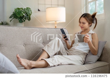 Relaxed Asian woman drinking coffee and using smartphone on couch. Concept of leisure and modern lifestyle 116196475