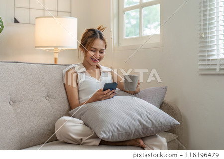 Young Woman Relaxing on Sofa with Smartphone and Coffee in Cozy Living Room Young Woman Relaxing on Sofa with Smartphone and Coffee in Cozy Living Room 116196479