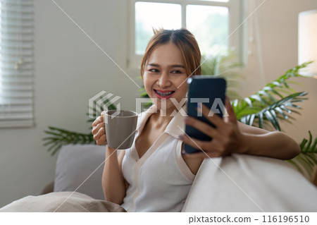 Young Woman Relaxing on Sofa with Coffee and Smartphone in Cozy Living Room Young Woman Relaxing on Sofa with Coffee and Smartphone in Cozy Living Room 116196510