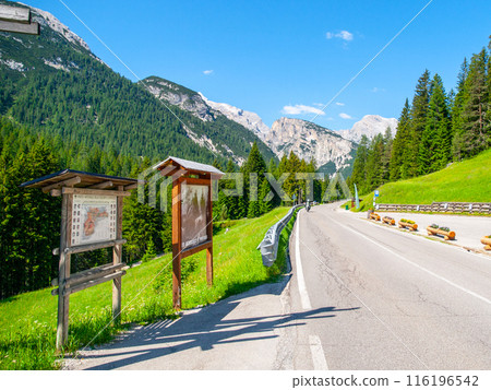 A winding road through the Italian Dolomites, with stunning mountain views and lush green trees. Two wooden signs with information stand on the side of the road. A winding road through the Italian Dolomites, with stunning mountain views and lush green trees. Two wooden signs with information stand on the side of the road. 116196542
