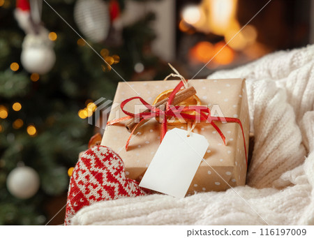 Gift tag on a Christmas present near sweater against Christmas tree lights and burning fire, mockup 116197009