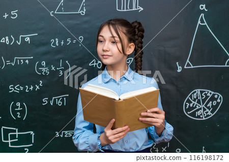 Beautiful teenage schoolgirl reading a textbook for a teacher while standing at a blackboard at school Beautiful teenage schoolgirl reading a textbook for a teacher while standing at a blackboard at school 116198172