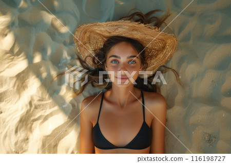 A beautiful girl lies in a black swimsuit on golden sand on a bright sunny day. Go-pro shot. top view, girl in a hat. distant perspective. The girl smiles. 116198727