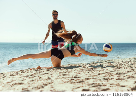 Focus on young woman playing beach volleyball with man, hitting ball in motion and falling down on sand. Dynamics and determination 116198804