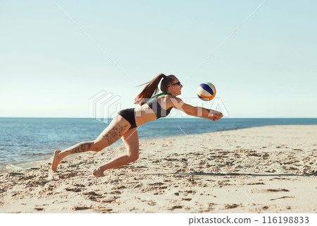 Concentrated and focused young girl in sportswear and sunglasses hitting ball and falling down, playing beach volleyball in the morning under clear sky Concentrated and focused young girl in sportswear and sunglasses hitting ball and falling down, playing beach volleyball in the morning under clear sky 116198833