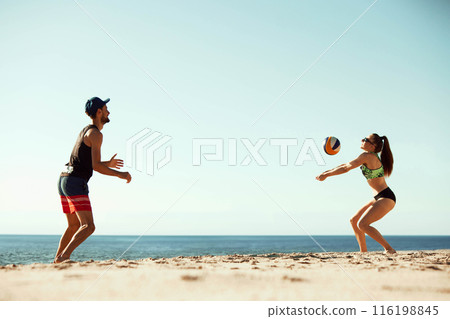 Volleyball players enjoying a sunny day at the beach, young man and woman playing beach volleyball under clear blue sunny sky Volleyball players enjoying a sunny day at the beach, young man and woman playing beach volleyball under clear blue sunny sky 116198845