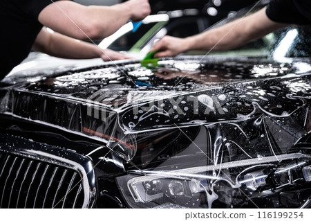 Mechanics applying PPF Paint Protection Film to a car hood in a detailing studio 116199254