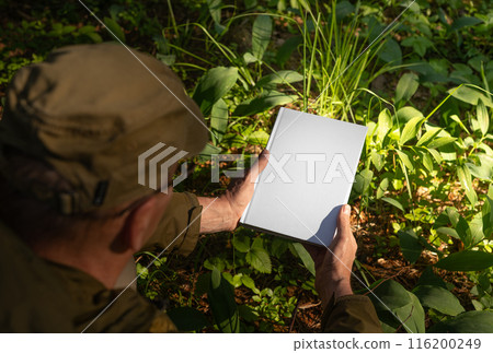 . Man holding book hard cover mockup, blank hardcover mock up in nature, green plants, grass, 116200249