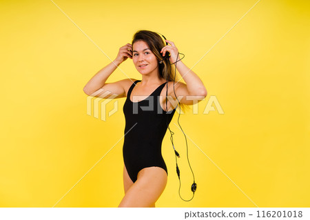 Caucasian Woman in Black Bodysuit Listening to Music in Headphones On Yellow Background. Caucasian Woman in Black Bodysuit Listening to Music in Headphones On Yellow Background. 116201018