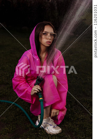 A glamour woman in a pink raincoat is spraying water from a hose 116201031