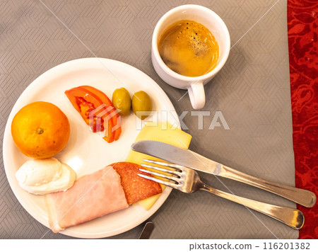 Hotel breakfast, orange, tomato slices, green olives, cheese slices, ham, salami, boiled egg, coffee, white plate Hotel breakfast, orange, tomato slices, green olives, cheese slices, ham, salami, boiled egg, coffee, white plate 116201382