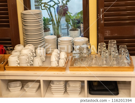 Hotel dining area, stacked plates, cups, glasses, trays, organized setup, breakfast service, white ceramic 116201394