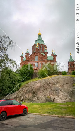 Uspensky Cathedral in Helsinki - red brick church with green blue roof on a cloudy day view from the parking with red car Uspensky Cathedral in Helsinki - red brick church with green blue roof on a cloudy day view from the parking with red car 116201950