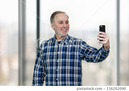 Portrait of smiling mature man taking a selfie on his smartphone camera. Standing indoors on windows backgorund. Portrait of smiling mature man taking a selfie on his smartphone camera. Standing indoors on windows backgorund. 116202090