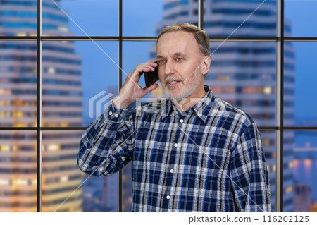 Handsome mature businessman is talking on the mobile phone standing in office. Checkered window background with view on the night city. Handsome mature businessman is talking on the mobile phone standing in office. Checkered window background with view on the night city. 116202125