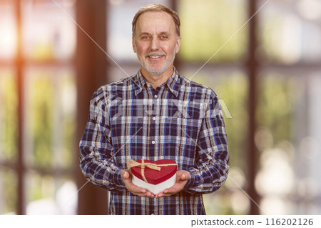Happy smiling handsome senior man is giving a heart shape gift box. Valentines day or anniversary. Blurred window background. Happy smiling handsome senior man is giving a heart shape gift box. Valentines day or anniversary. Blurred window background. 116202126