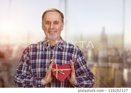 Happy smiling handsome senior man is holding a heart shape gift box. Blurred cityscape background. Happy smiling handsome senior man is holding a heart shape gift box. Blurred cityscape background. 116202127