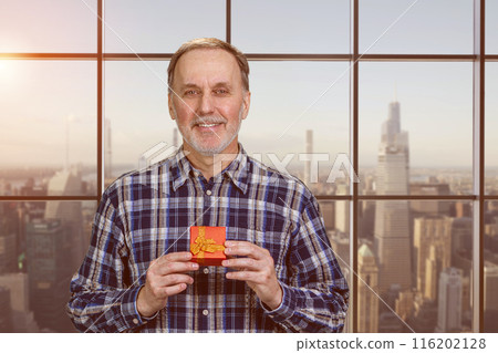 Happy smiling handsome senior man is holding a red gift box in both hands. Evening cityscape background. Happy smiling handsome senior man is holding a red gift box in both hands. Evening cityscape background. 116202128