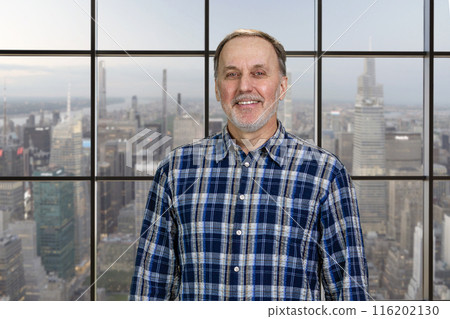 Portrait of smiling cheerful mature senior man standing indoors. Checkered windows background with cityscape view. 116202130