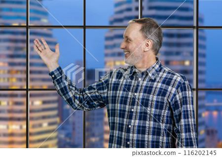Handsome cheerful mature senior man presenting copy space with his hand. Checkered windows with night cityscape background. 116202145