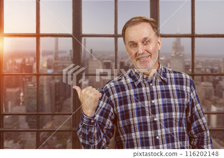 Portrait of smiling mature man pointing back with his thumb up. Checkered windows background with cityscape view. Portrait of smiling mature man pointing back with his thumb up. Checkered windows background with cityscape view. 116202148