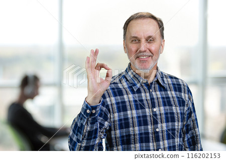 Smiling confident mature man giving an okay gesture. Office enviroment background. Smiling confident mature man giving an okay gesture. Office enviroment background. 116202153