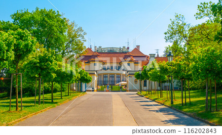 A paved driveway leads to the entrance of the Kramars Villa, the official residence of the Czech Prime Minister in Prague. Lush green trees line the path, providing shade on a sunny day. 116202843
