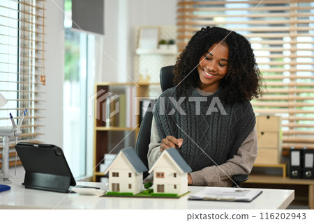 Smiling African American woman with house model and wind turbine, sustainable energy and modern technology 116202943