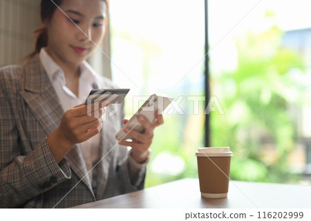 Young businesswoman holding a credit card making online payment on mobile phone, sitting at workplace table Young businesswoman holding a credit card making online payment on mobile phone, sitting at workplace table 116202999