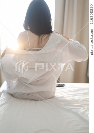 Young woman in lingerie and white shirt sit on bed indoors at home stretching in the morning after waking up, back view. Relaxing , Cozy, good morning, comfort, intimacy concept Young woman in lingerie and white shirt sit on bed indoors at home stretching in the morning after waking up, back view. Relaxing , Cozy, good morning, comfort, intimacy concept 116208830