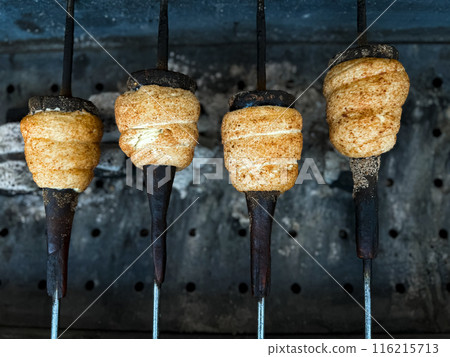 Trdelnik baking on metal rods over hot coals. Close up view of traditional Hungarian street food being prepared at market. Food and culinary concept for poster and print Trdelnik baking on metal rods over hot coals. Close up view of traditional Hungarian street food being prepared at market. Food and culinary concept for poster and print 116215713