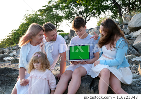 A family of five sits with a laptop in the mountains near the ocean Cheerful family with kids on beautiful summer meadow learning on laptop Family laptop green screen A family of five sits with a laptop in the mountains near the ocean Cheerful family with kids on beautiful summer meadow learning on laptop Family laptop green screen 116223348
