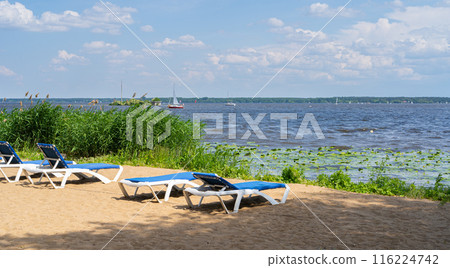 Sunbeds on lake sand beach, blue lounge chairs, deck chairs near water in reeds, boat docked, blue sky Sunbeds on lake sand beach, blue lounge chairs, deck chairs near water in reeds, boat docked, blue sky 116224742
