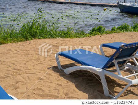 Sunbeds on lake sand beach, blue lounge chairs, deck chairs near water in reeds, boat docked, blue sky Sunbeds on lake sand beach, blue lounge chairs, deck chairs near water in reeds, boat docked, blue sky 116224743