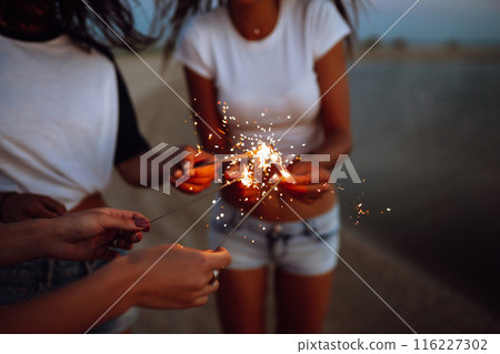 The sparklers in the hands of young girls on the beach. Three girls enjoying party on beach with sparklers. 116227302