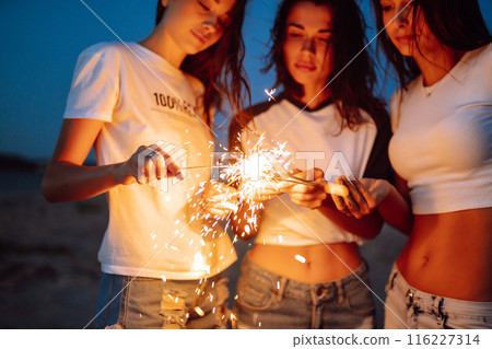 The sparklers in the hands of young girls on the beach. Three girls enjoying party on beach with sparklers. The sparklers in the hands of young girls on the beach. Three girls enjoying party on beach with sparklers. 116227314