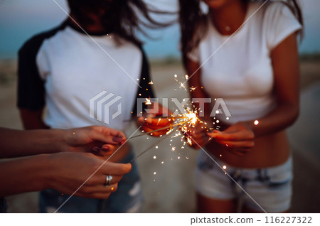 The sparklers in the hands of young girls on the beach. Three girls enjoying party on beach with sparklers. The sparklers in the hands of young girls on the beach. Three girls enjoying party on beach with sparklers. 116227322