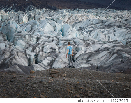 A man stands on a rocky outcropping, dwarfed by a vast glacier in Iceland. The glacier's crevasses and ice formations stretch out as far as the eye can see. 116227665
