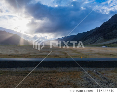 A paved road cuts through a vast, flat plain in Iceland, with mountains in the distance. The cloudy sky and sunlight create dramatic lighting and long shadows. A paved road cuts through a vast, flat plain in Iceland, with mountains in the distance. The cloudy sky and sunlight create dramatic lighting and long shadows. 116227710